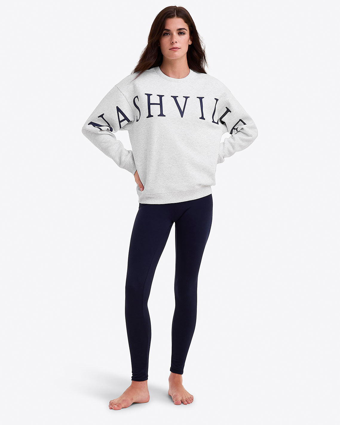 A woman stands barefoot in a Draper James Nashville Crewneck Sweatshirt and black leggings against a white background, embodying effortless country style.