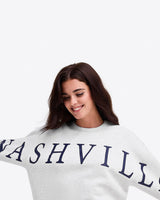 Woman with long brown hair smiling and wearing the Draper James Nashville Crewneck Sweatshirt in light gray with bold navy lettering—ideal for country music fans.