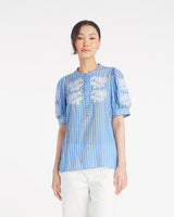 A woman models the Draper James Puff Sleeve Top with Floral Embroideryâ€”featuring blue and white stripesâ€”styled with white pants against a plain background, perfect for effortless summer style.