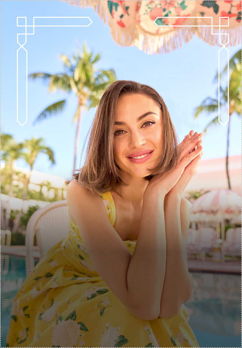 Woman in a yellow dress with floral patterns by a poolside with palm trees in the background