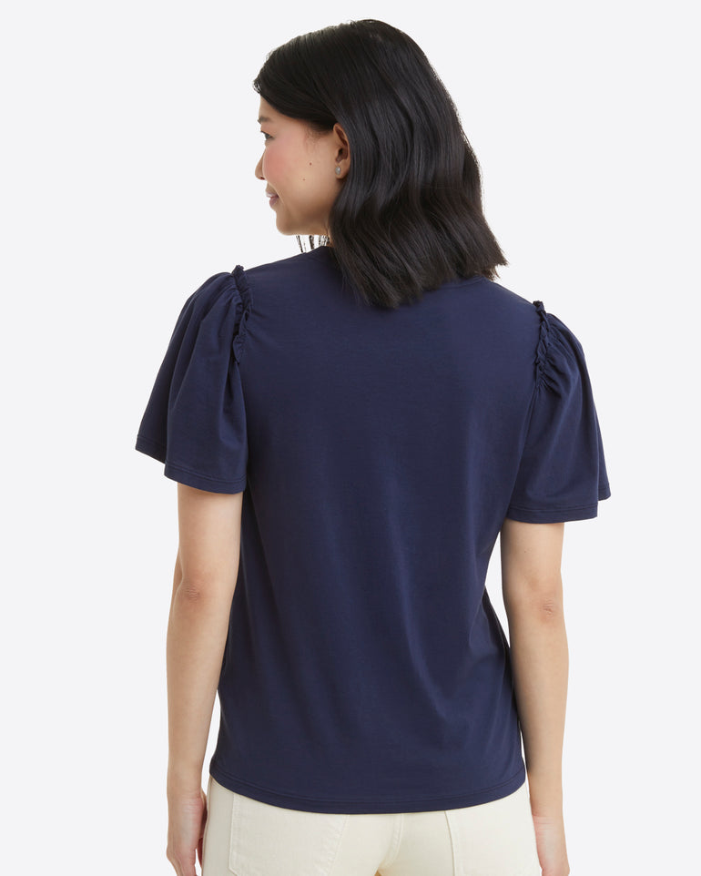 A person with shoulder-length hair, wearing the Draper James Short Sleeve Easy Knit Top in Navy made from 100% Pima Cotton and light-colored pants, is facing away from the camera against a white background.
