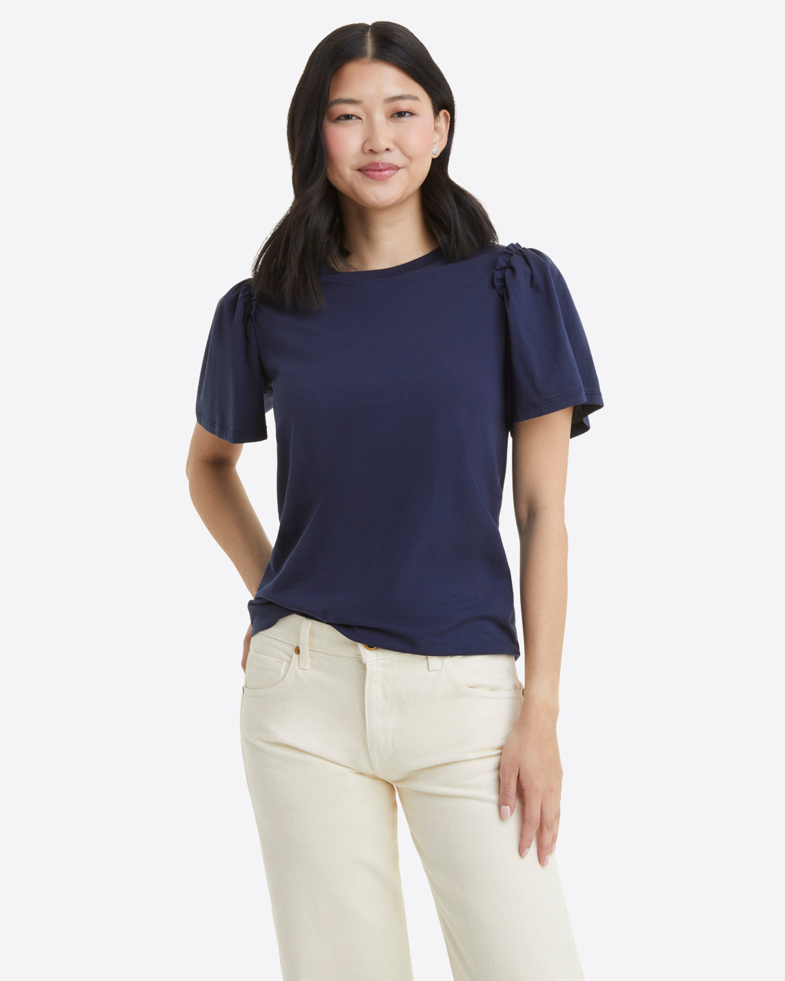A woman stands against a plain background, slightly smiling as she faces forward. She wears a Draper James Short Sleeve Easy Knit Top in Navy, made from 100% Pima Cotton, paired with light-colored pants.