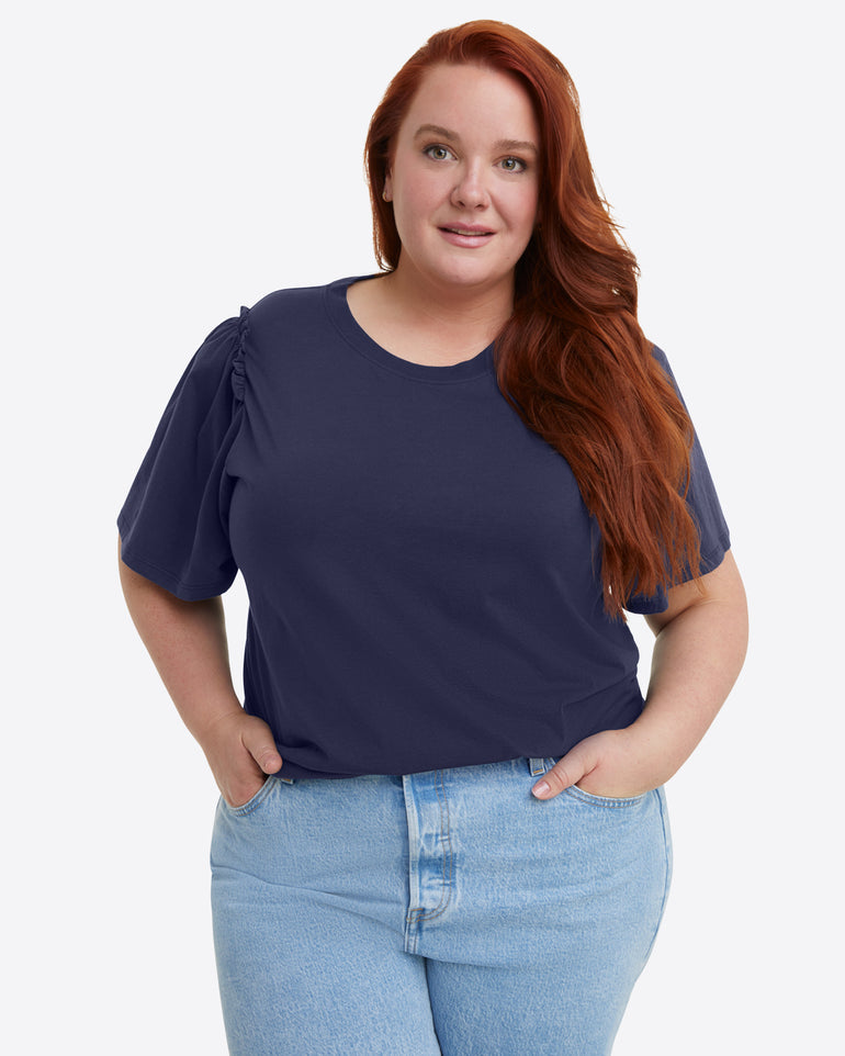 A person with long red hair is wearing a Draper James Short Sleeve Easy Knit Top in Navy made from 100% Pima Cotton, paired with light blue jeans, against a plain background.